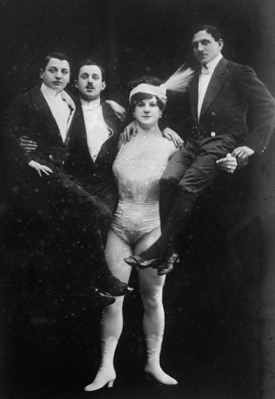 Es ist eine Frau zu erkennen mit stämmigen Körperbau, die links einen Mann und rechts 2 Männer auf den Armen in die Höhe hält.