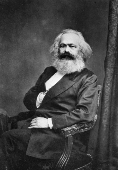 Karl Marx, er trägt einen Anzug, die rechte Hand hat er unter dem Revers die linke Hand ruht auf seinem linken Oberschenkel. Karl Marx hat langes graues Haar und einen grauen Vollbart und eien schwarzen ausgeprägten Schnurrbart.