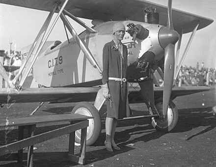 Auf dem schwarz-weiss Foto ist eine Frau, um die 30 Jahre alt, vor einem Propellerflugzeug zu sehen. Im Hintergrund ist eine Zuschauertribüne mit vielen Menschen.  Das Foto entstand um 1928.