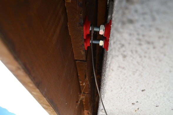 Zwei Klemmen sind nebeneinander im montiertem Zustand zu sehen. An der näheren Balkonklemme hängt ein 2 Millimeter starkes Stahlseil um die Schraube, das nach unten weghängt.