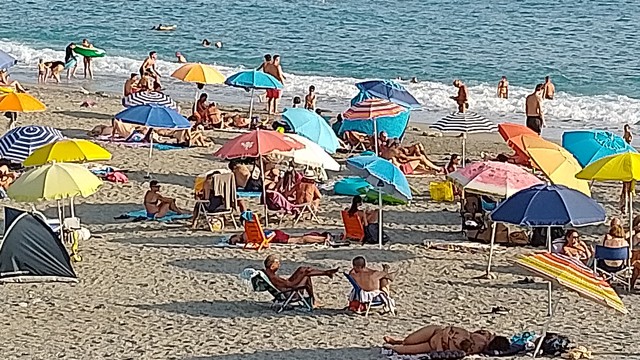Menschen am Strand unter Sonnenschirmen, dahinter das Meer mit weißen Schaumkronen auf den Brandungswellen.