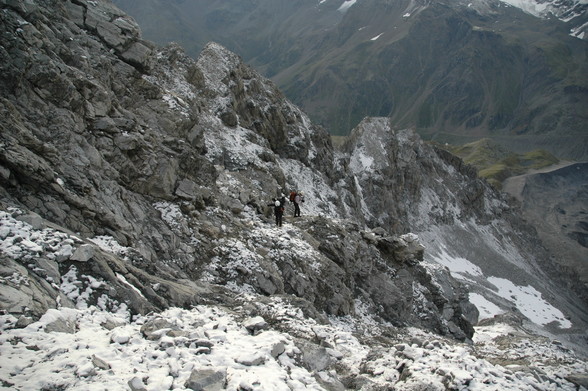 Zwei Alpinisten im hochalpinen Gelände am Ortler Hintergrad.
Beide tragen einen weißen Helm und einen großen Rucksack.
Das Gelände ist relativ steil und zerklüftet, nach rechts in abfallend. Es liegt einige Centimeter Schnee auf den losen Steinen.