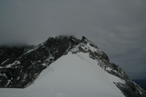 Ein größeres Schneefeld im Vordergrund, welches zuerst etwas abfällt und dann wieder ansteigt und dann übergeht in leicht verschneites Stein-/Felsengelände. Man sieht ein paar Bergsteiger etwas nach der Senke, und ein paar Richtung Ende des Schneefeldes, die sind entfernungsbedingt schon sehr klein.
Bewölktes Wetter, die Wolken liegen schon etwas tiefer als die höchsten Stellen des Hintergrates.