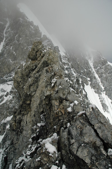 Scharfer/spitzer schmaler Berggrat im Vordergrund, im Hintergrund sieht man ein steiles Schneefeld über das die Route raufführt.
Ganz links unten in der Bildecke sieht man einen Bergsteiger der an den Felsen entlangklettert.
Das Schneefeld im Hintergrund liegt schon im leichten Nebel.