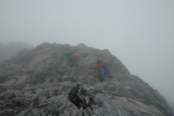 Zwei Bergsteiger die mit Steigeisen eine steile Felspassage raufklettern. Der vordere hat es fast geschafft.
Die Bergsteiger sind von einem Seil, das von oben herabläuft, gesichert. Der Rest dieser Gruppe und der Sichernde sind hinter dem am Bild sichtbaren höchsten Punkt.
Etwas Nebel trübt das Bild.